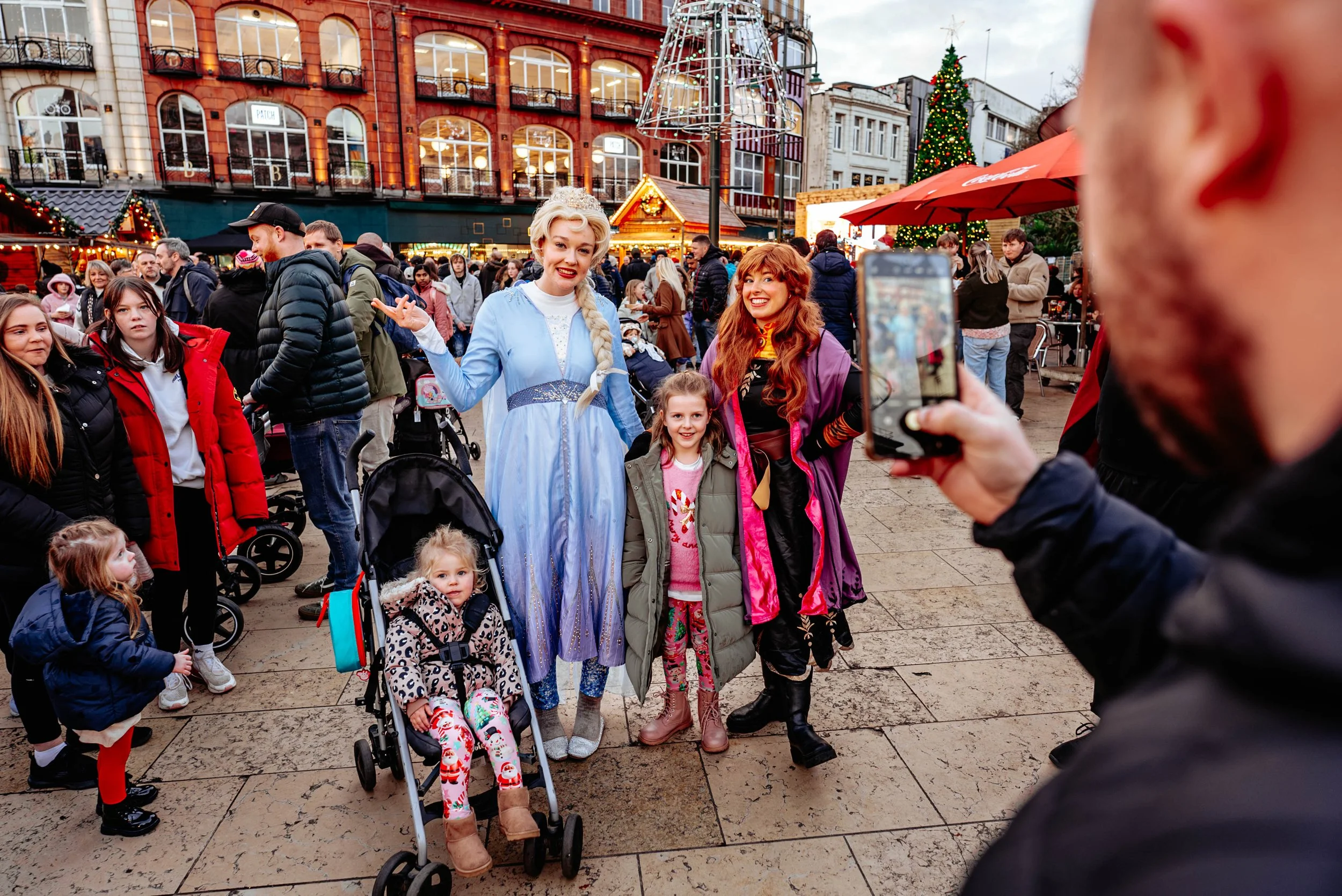 People dressed as Elsa and Anna from Frozen taking a photo with a young girl at a festive outdoor holiday market with Christmas decorations and a decorated tree in the background.