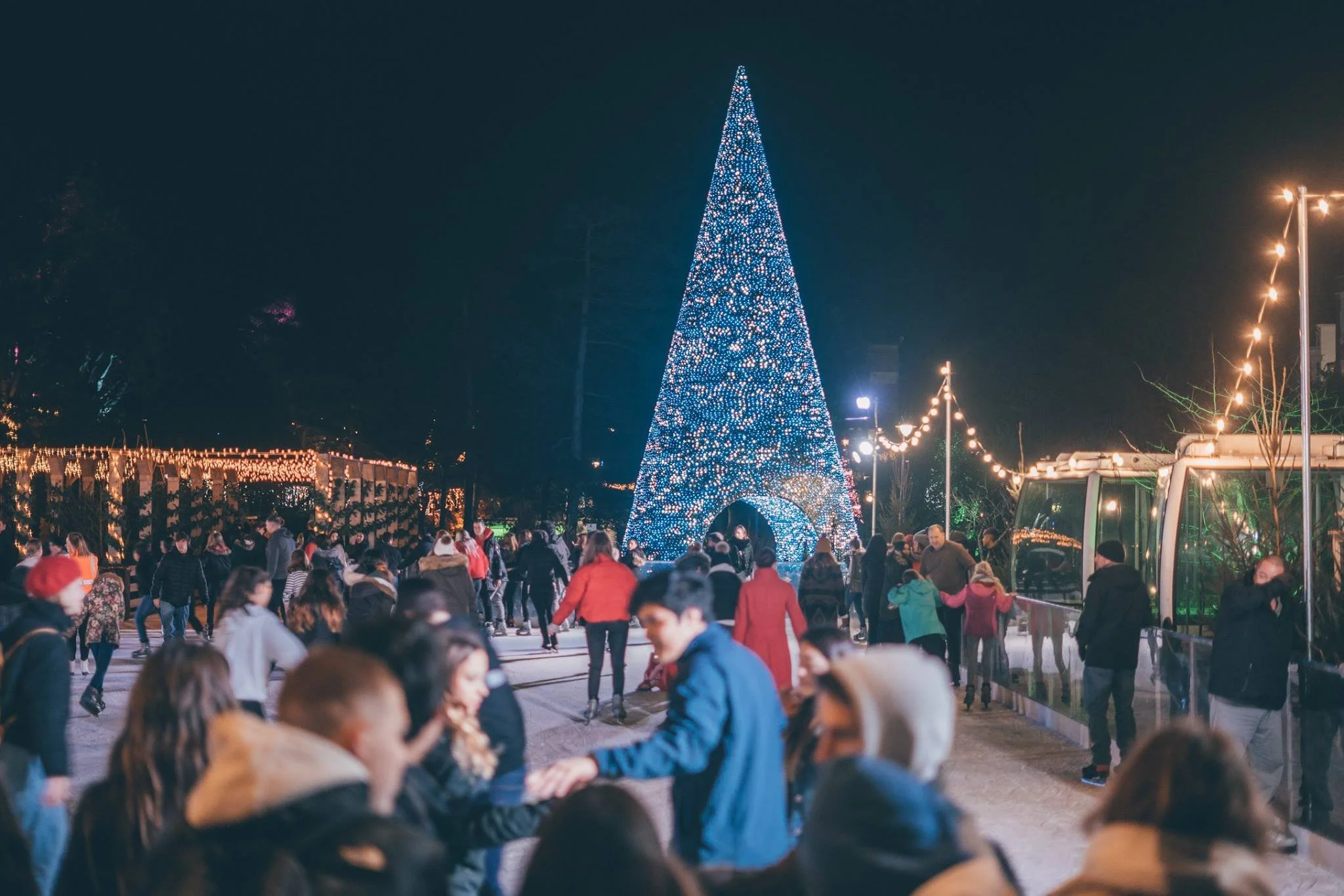 People skating outdoors at night near a large, illuminated Christmas tree decorated with blue and white lights.