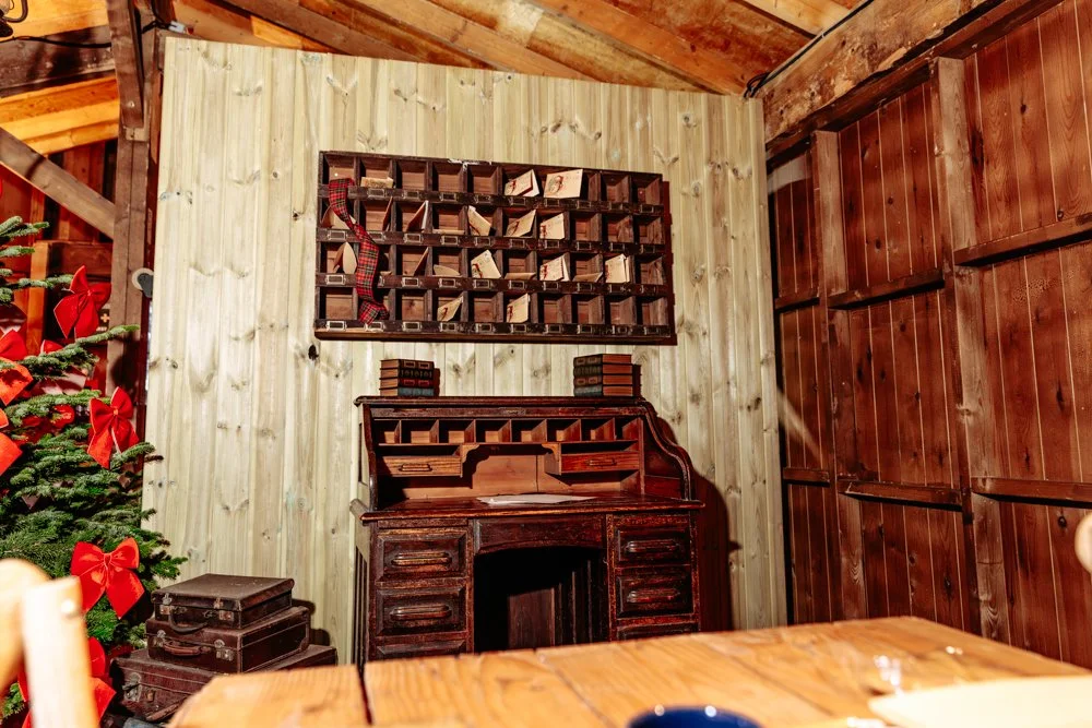 A Christmas tree decorated with red bows on the left, a wooden desk with old books on top, a wall-mounted mail sorting unit, and wood-paneled walls and ceiling in a rustic room.