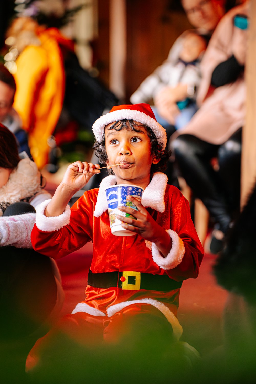 Young boy dressed as Santa Claus sipping from a cup at a festive gathering with others in the background.