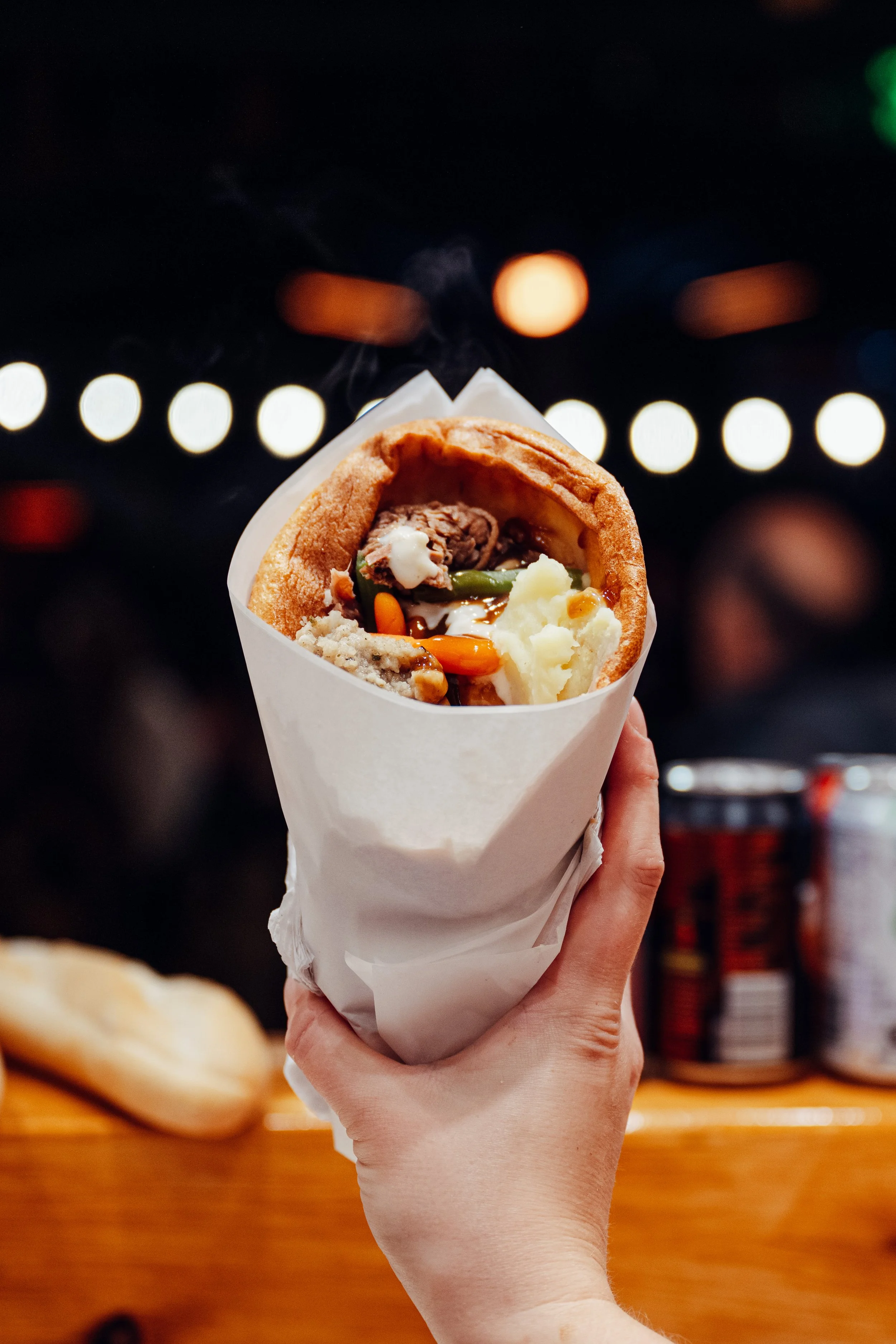 A hand holding a steaming wrap with beef, mashed potatoes, vegetables, and gravy, in a paper wrapper, against a blurred background with bokeh lights.