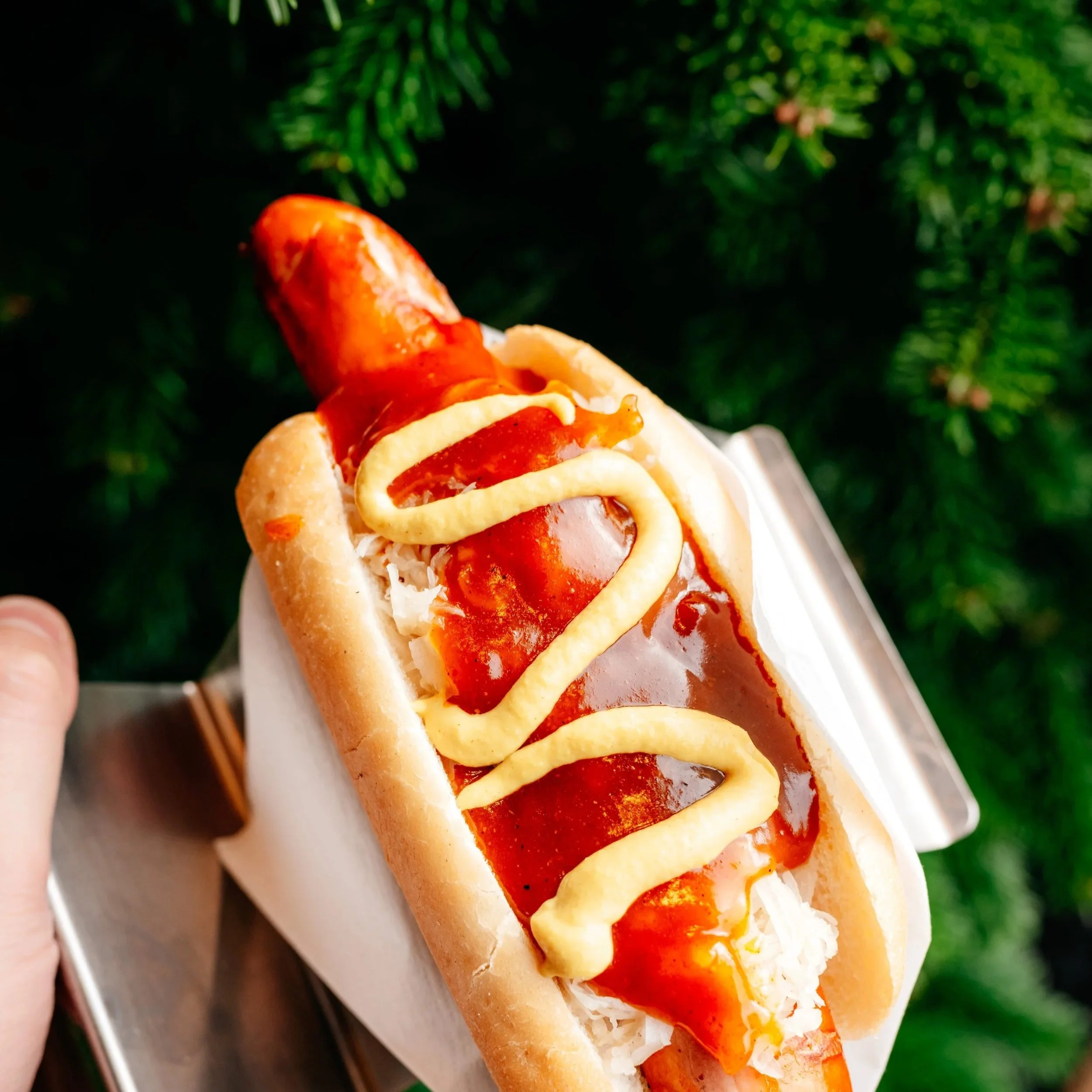 Hot dog in a bun topped with mustard, ketchup, and shredded cheese, held outdoors with green foliage in the background.