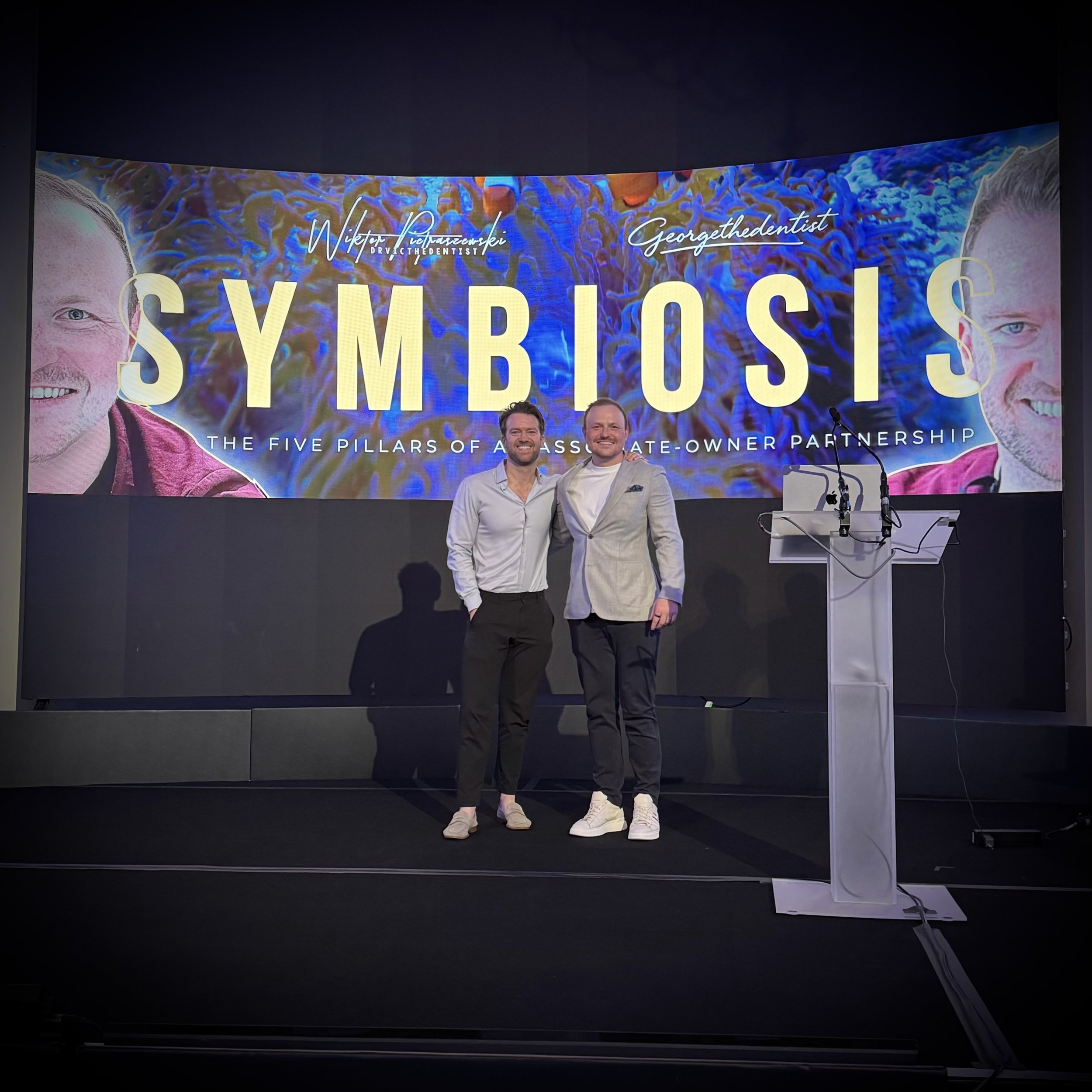 Two men standing on a stage in front of a large screen displaying the word "SYMBOLS" and the subtitle "The five pillars of a state owner partnership." Both are smiling, one wearing a white shirt and black pants, the other wearing a light gray jacket,