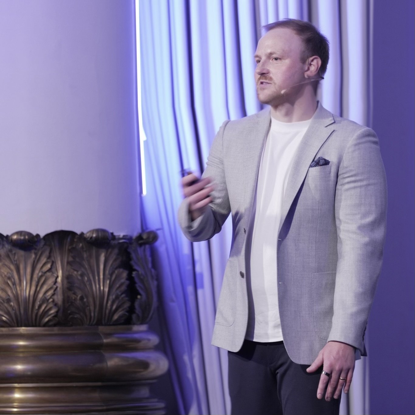 A man in a light gray blazer and white shirt giving a presentation or speech in front of a curtain with blue lighting.