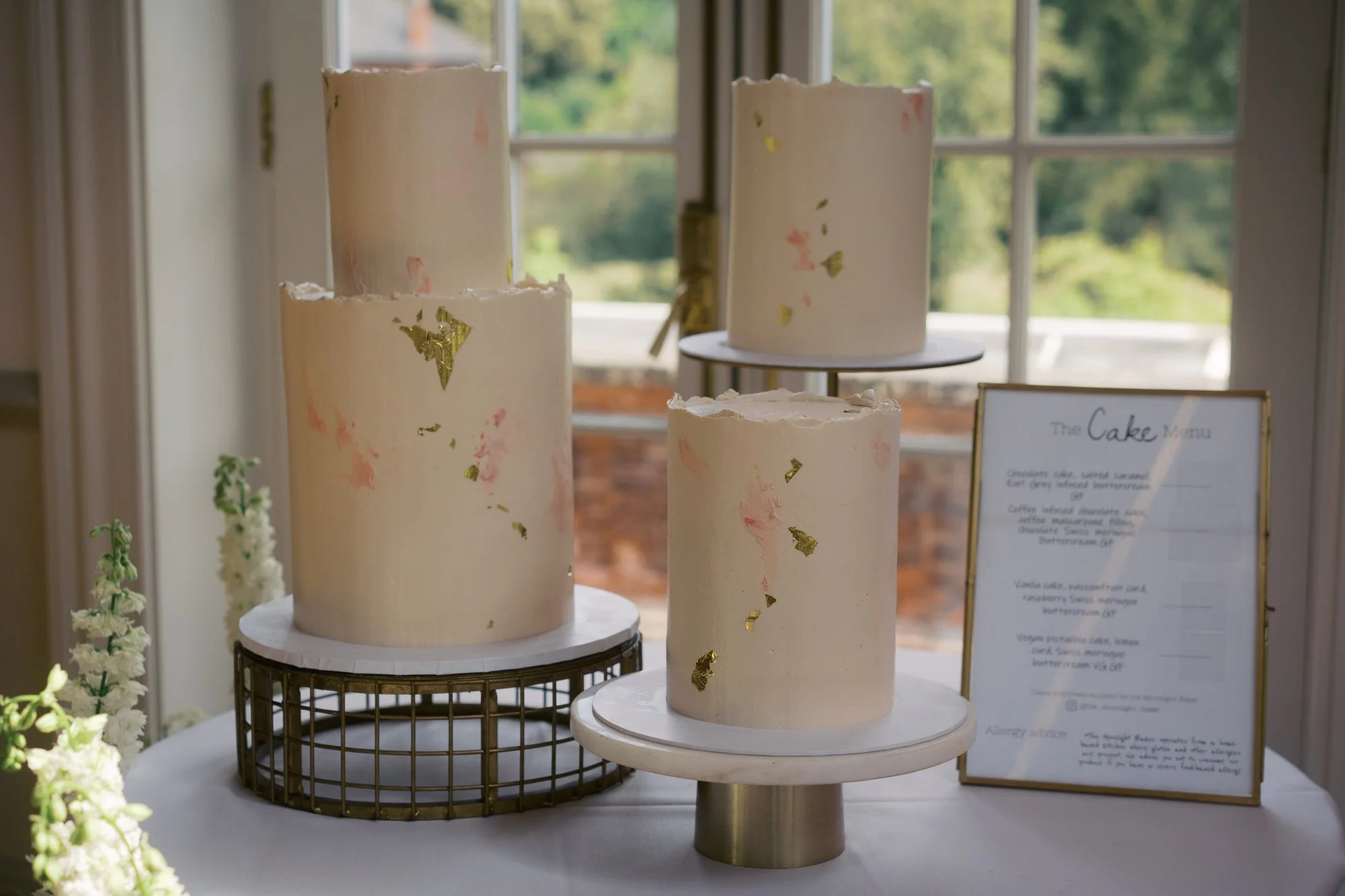 A white wedding cake with gold leaf by a window at Hodsock Priory