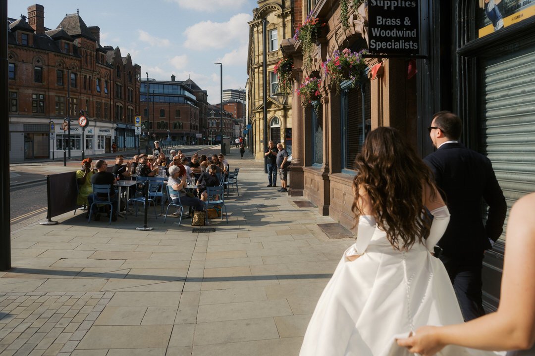  Bride and groom walking along a sunny Leeds street as people at the tables outside the Adelphi pub turn to watch. 
