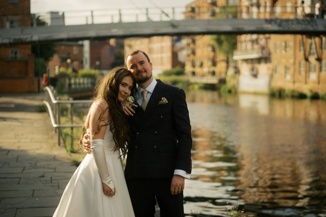  Bride and groom leaning into each other beside the water in Leeds in the evening sun, with a footbridge and old warehouses behind them. 