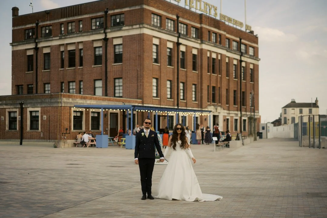  Bride and groom in sunglasses holding hands on the square in front of The Tetley in Leeds, with guests on the terrace behind them. 