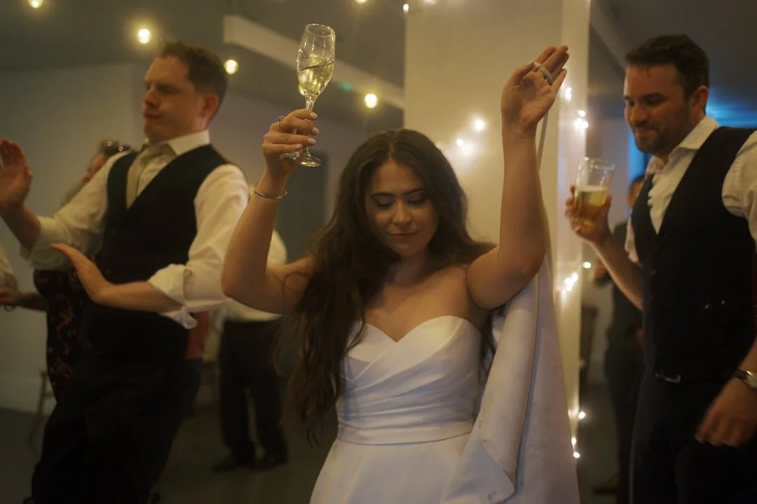 Bride dancing with her arms raised and a glass of fizz in one hand, among guests on the dance floor at The Tetley in Leeds. 