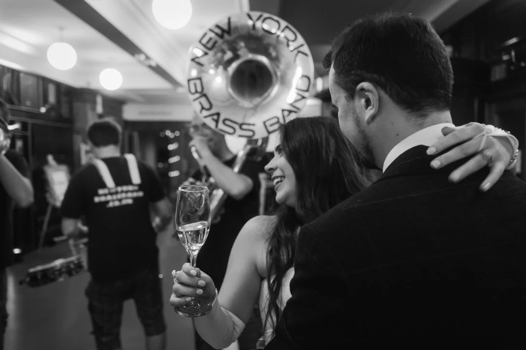  Black and white photo of the bride and groom embracing, the bride holding a glass of fizz, as the New York Brass Band plays behind them at The Tetley in Leeds. 