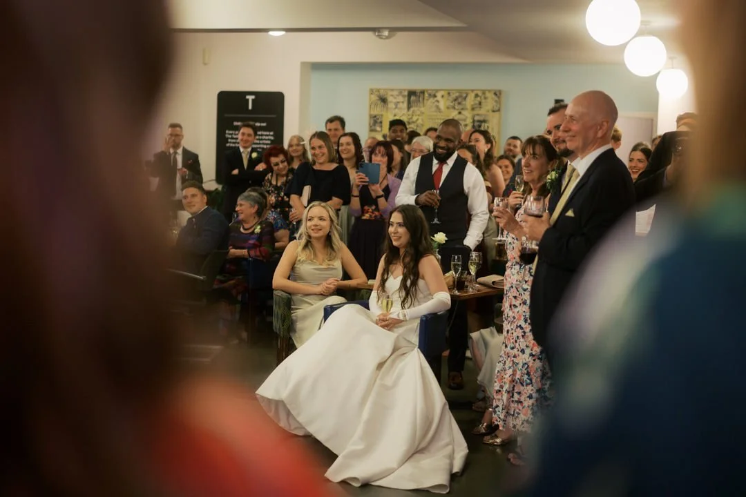  Bride laughing during the speeches at The Tetley in Leeds, seated beside a bridesmaid with guests standing around them. 