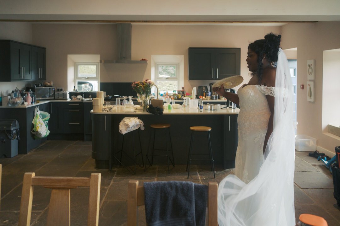  The bride in her dress and veil with a woven fan, in the barn kitchen 