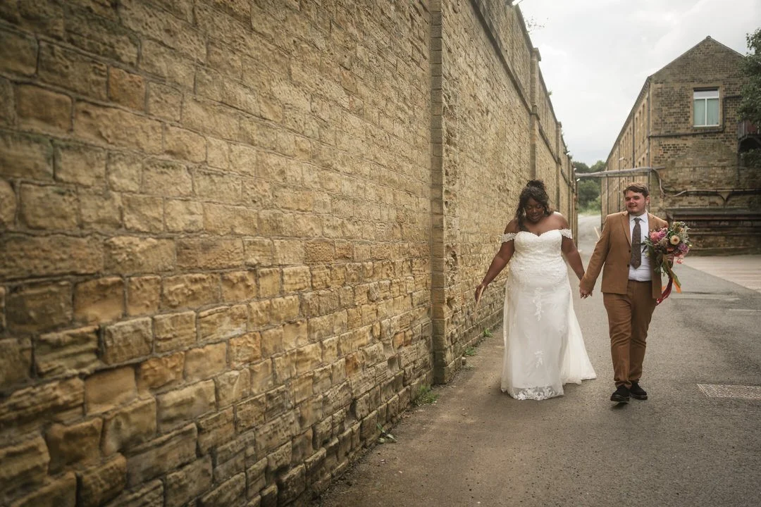  The bride and groom walking beside a tall sandstone mill wall at Bowers Mill, near Halifax 