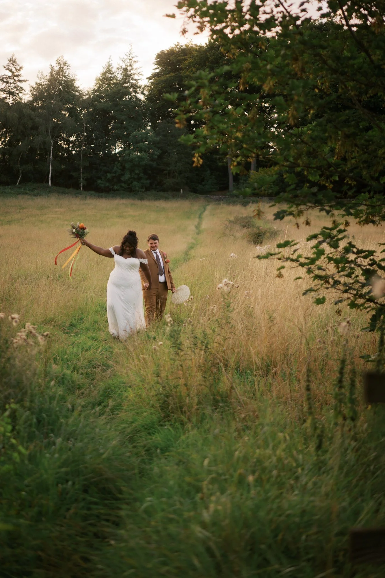  The bride and groom walking through long grass in the evening, the bride holding her bouquet up 