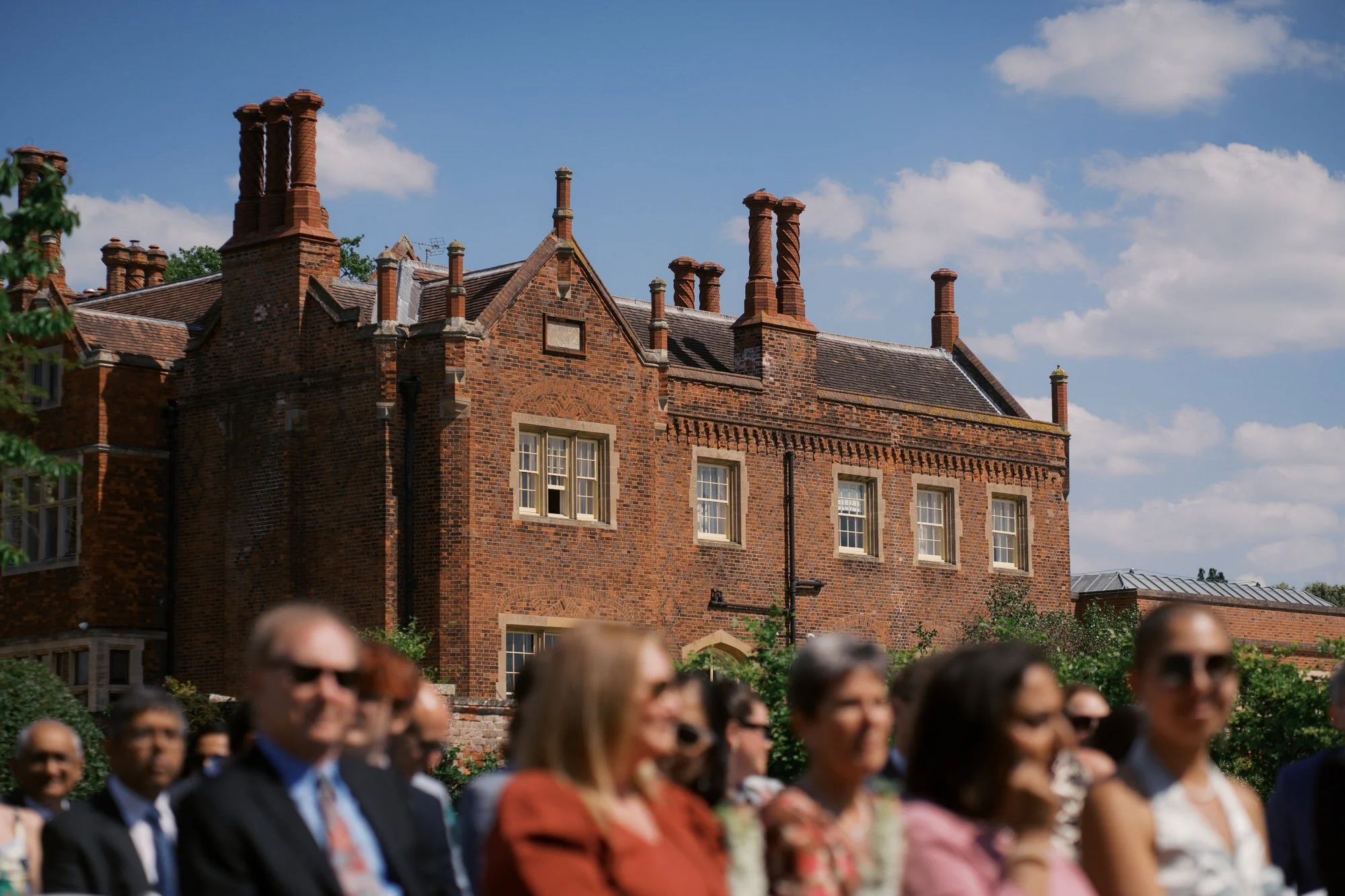  Seated wedding guests in the foreground with the red-brick Hodsock Priory in focus behind them 