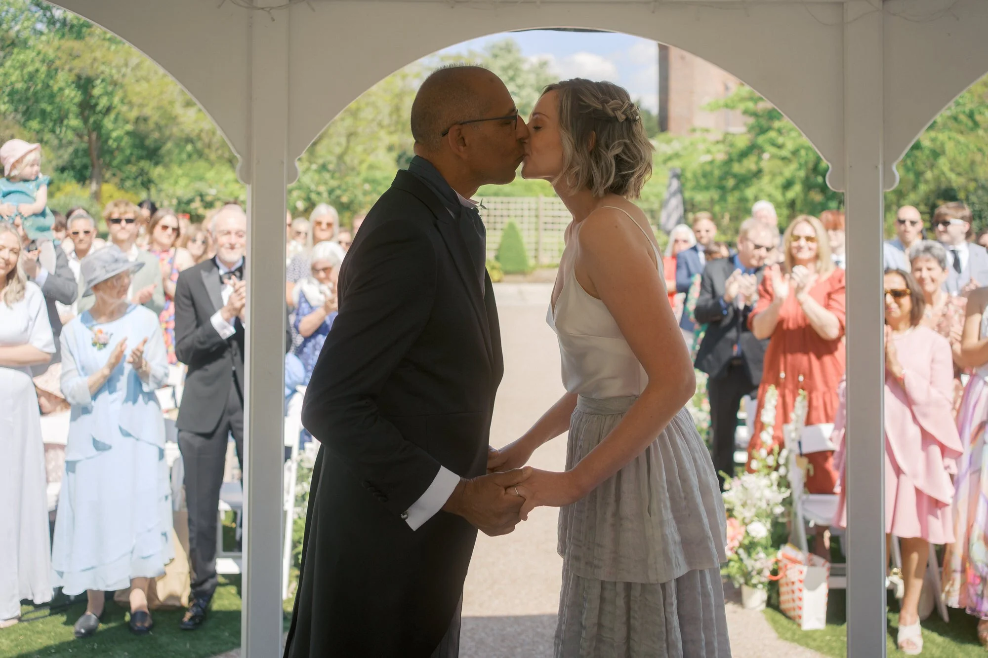  A couple kissing beneath a white garden pavilion as guests applaud 