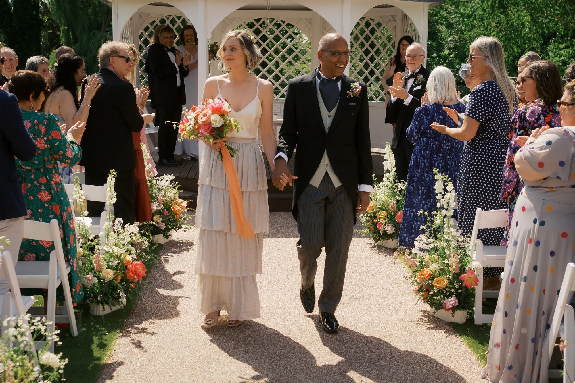  A newly married couple walking back up a garden aisle, smiling, as guests applaud 