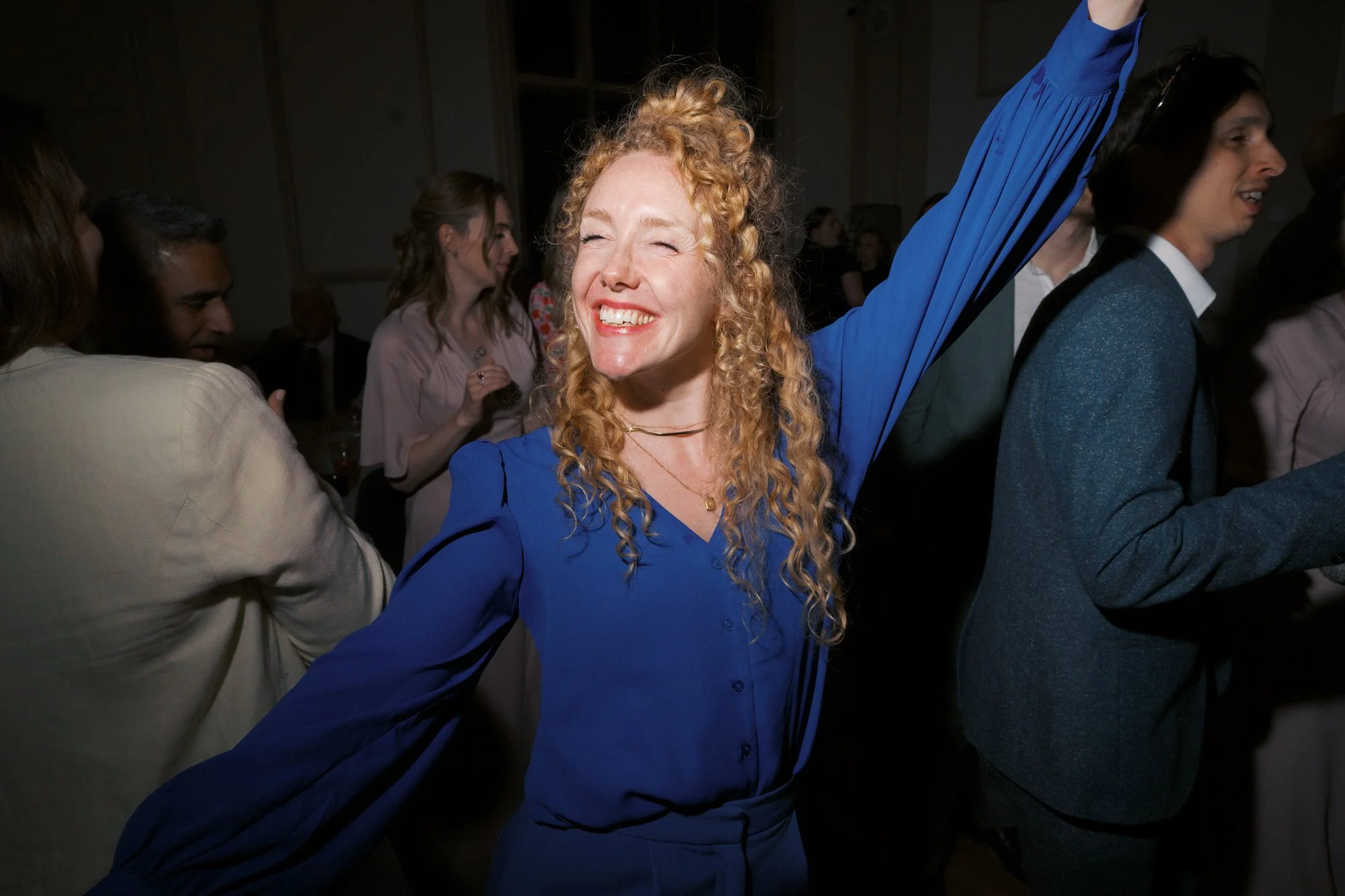 A wedding guest in blue dancing with one arm in the air on a busy dance floor 