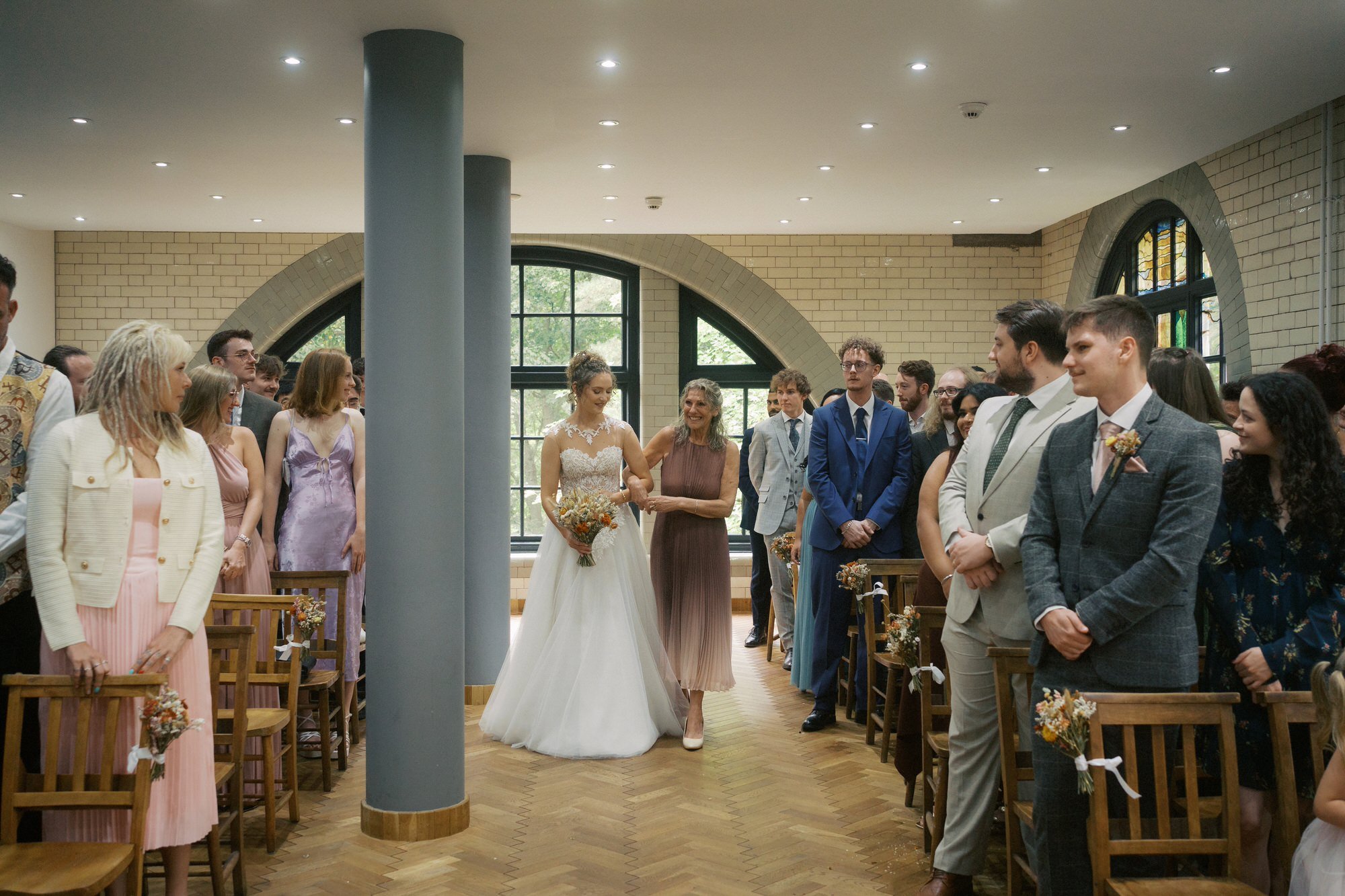  A bride walking down the aisle arm in arm with a woman in a pleated dress as guests turn to watch, The Pumping House, Ollerton 