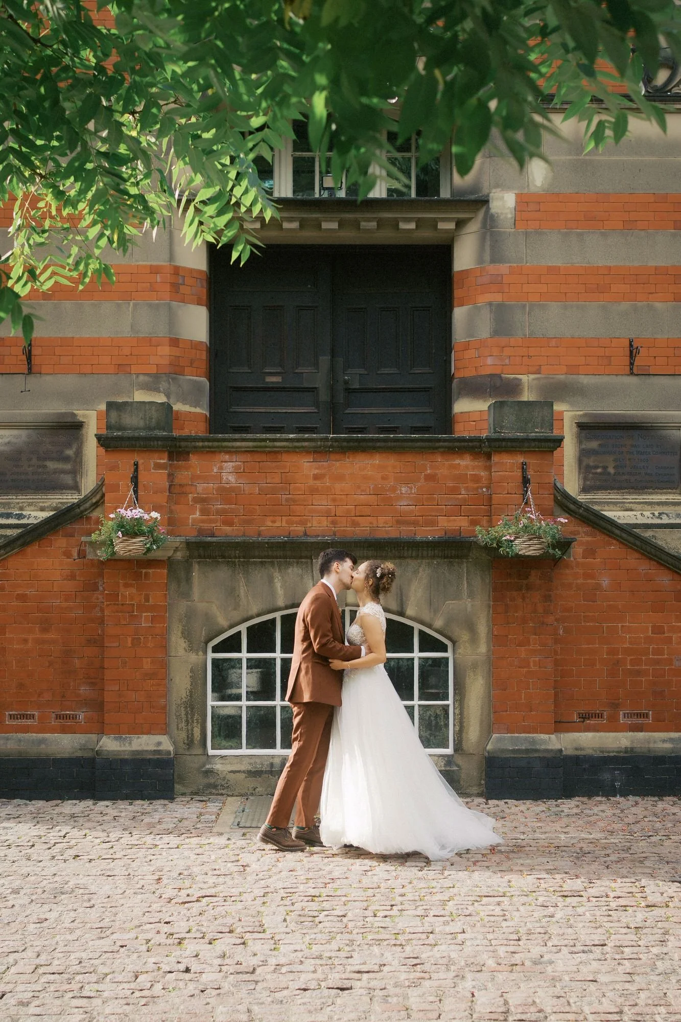  A groom and bride kissing on the cobbles in front of the striped red-brick entrance of The Pumping House, Ollerton 