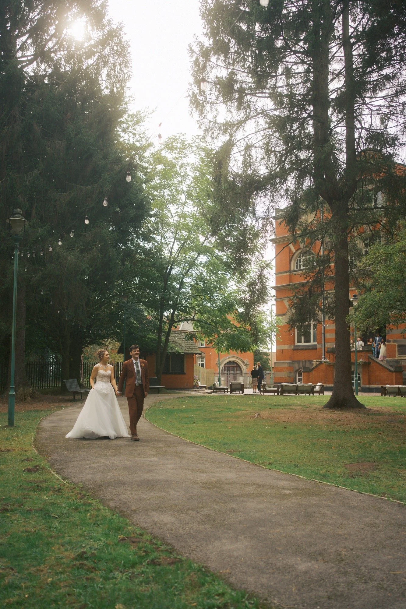  A bride and groom holding hands on a path in low sun through the trees, The Pumping House, Ollerton 