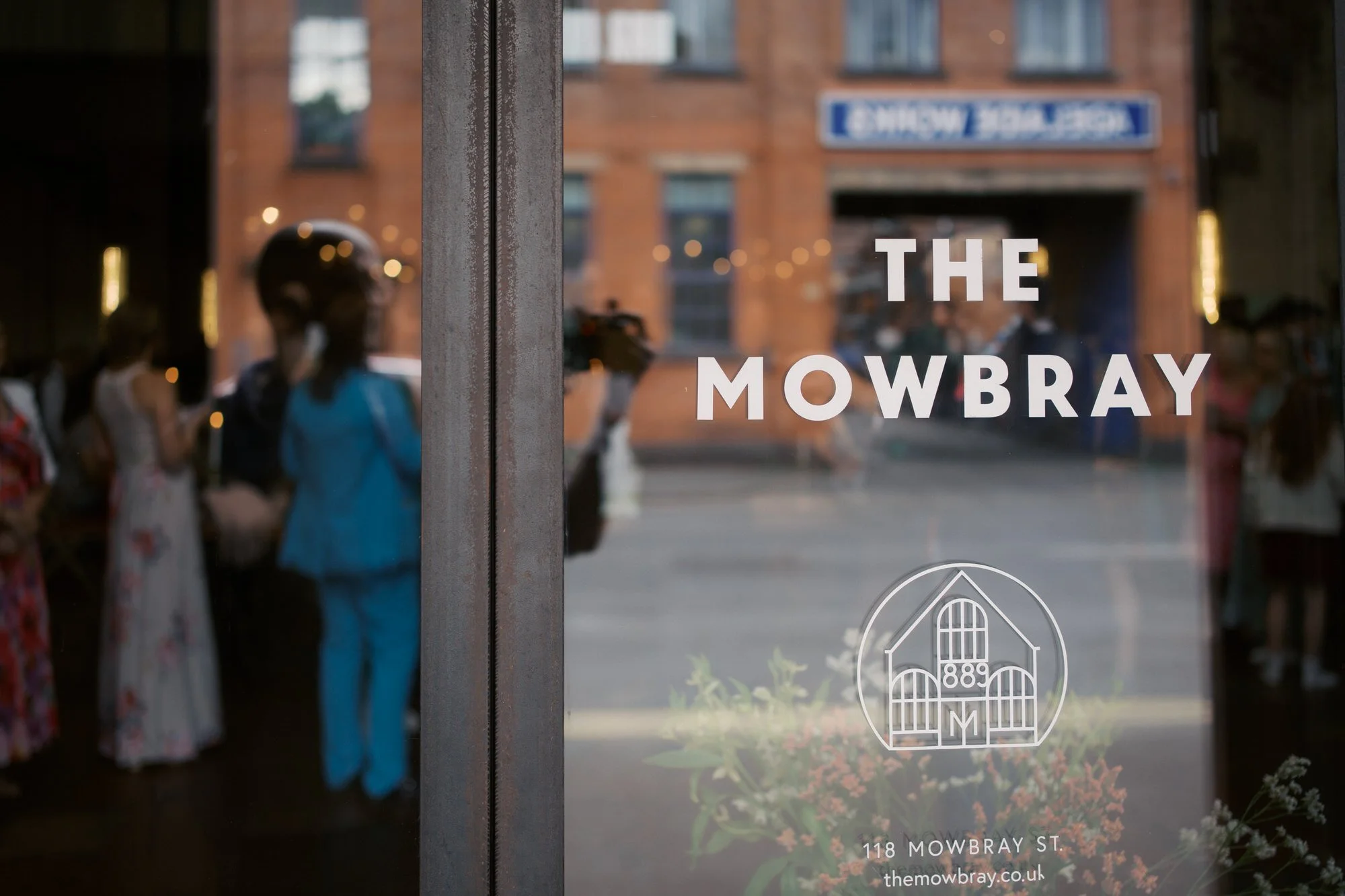 Wedding guests gathered inside the entrance of The Mowbray in Neepsend, Sheffield, seen through the glass door with the venue's name on it