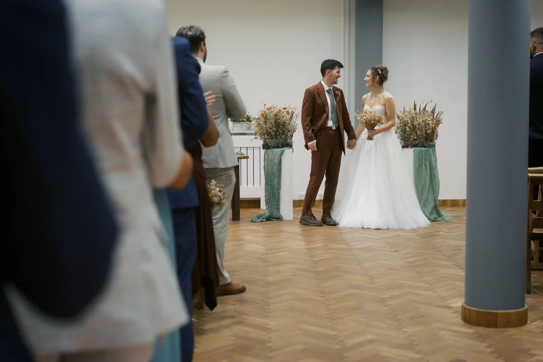 Bride and groom holding hands and smiling at each other at the front of the ceremony room at The Pumping House, between two arrangements of dried flowers, with standing guests in the foreground.