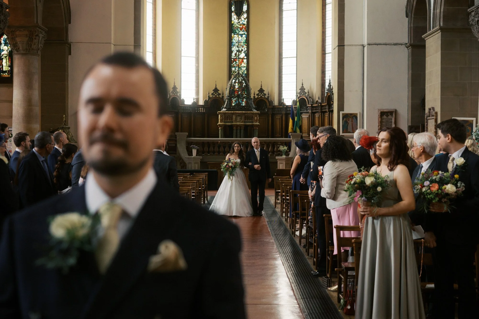 Bride walking down the aisle of St Aidan's Church in Harehills on her dad's arm, while the groom waits in the foreground with his eyes closed.
