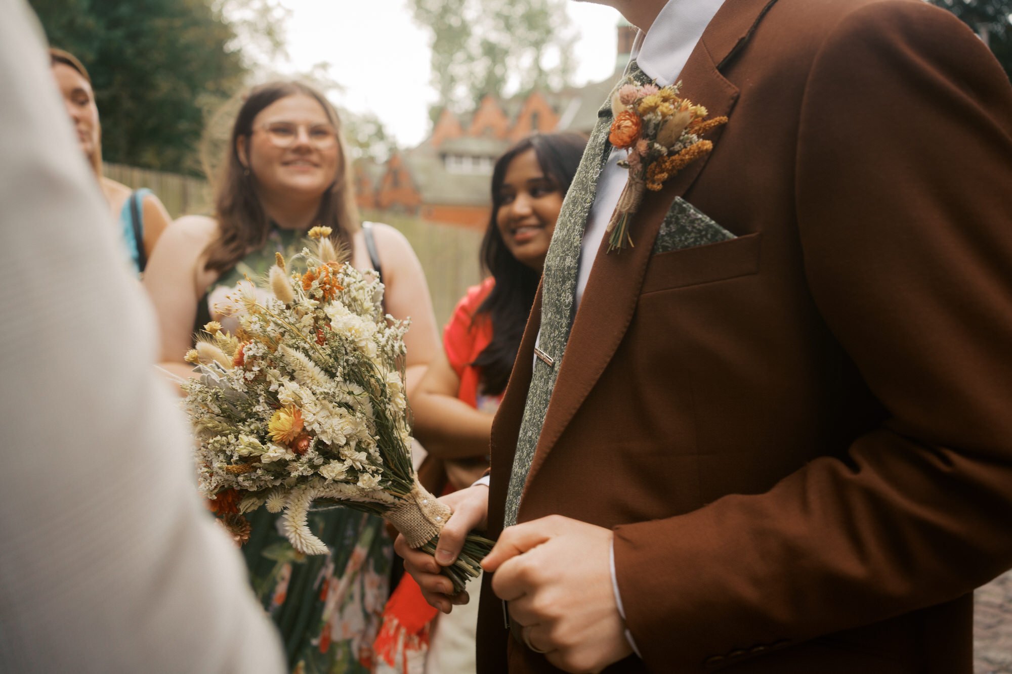 Groom in a rust-brown jacket holds a dried flower bouquet while smiling guests chat around him outdoors