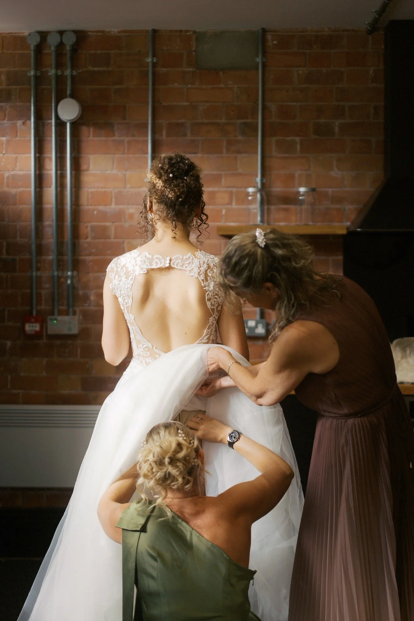 A bride's mother and another woman helping with the back of her wedding dress against an exposed brick wall, The Pumping House Quarters, Ollerton
