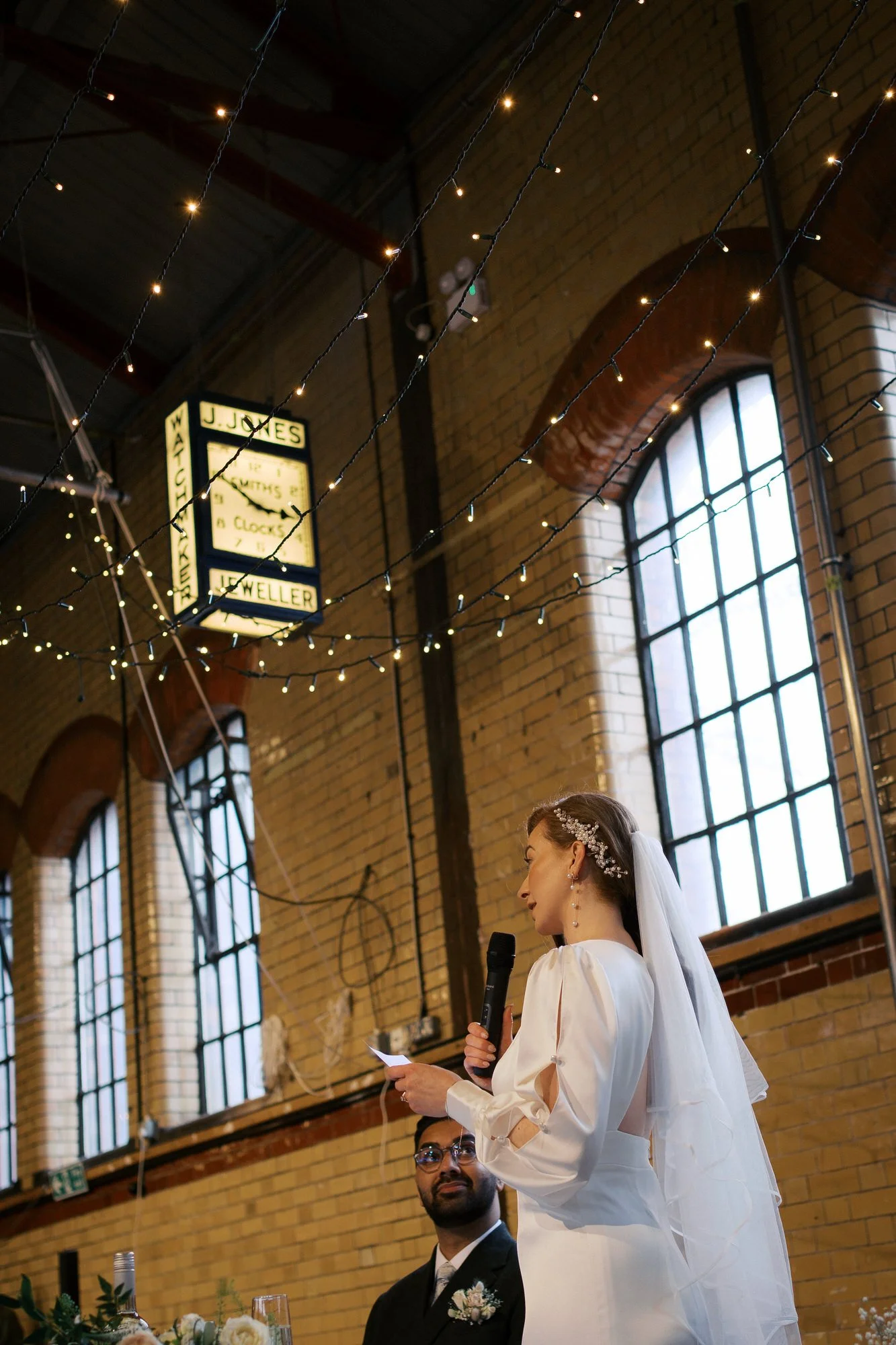 bride giving her speech beneath a vintage illuminated watchmaker's clock and strings of fairy lights in a tiled industrial hall at Kelham Island Museum, Sheffield
