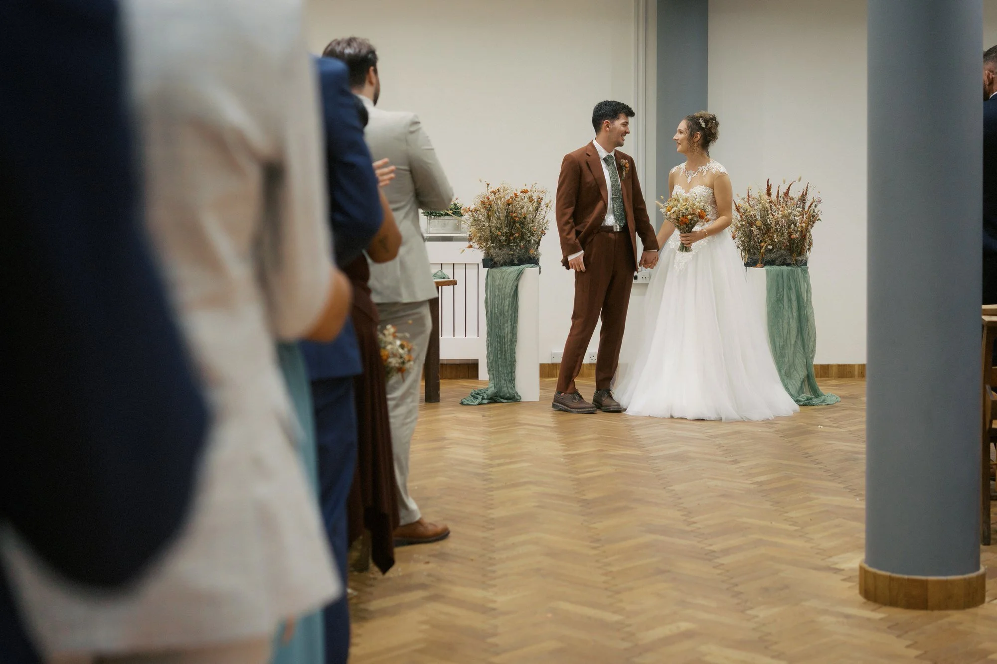 A bride and groom holding hands and smiling at each other, seen between guests along the aisle, The Pumping House, Ollerton