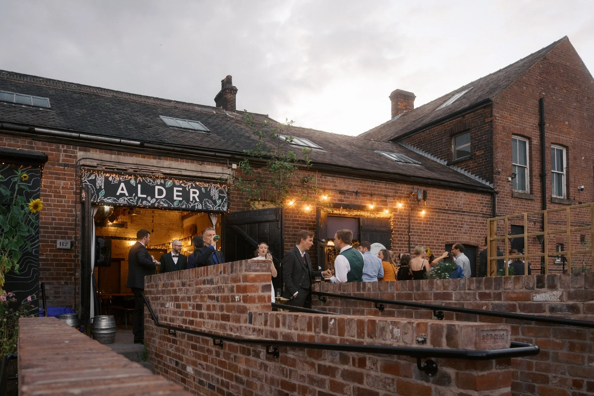 Wedding guests chatting with drinks in the brick courtyard outside Alder on Percy Street, Neepsend, Sheffield, at dusk