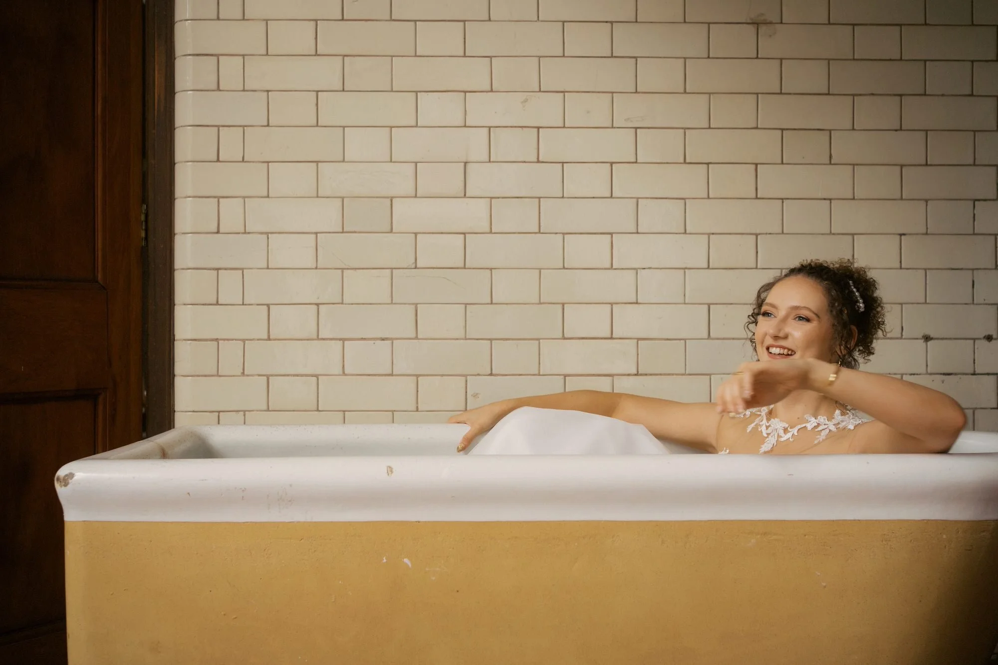 A bride in her wedding dress laughing as she reclines in an old tub against tiled walls, The Pumping House, Ollerton