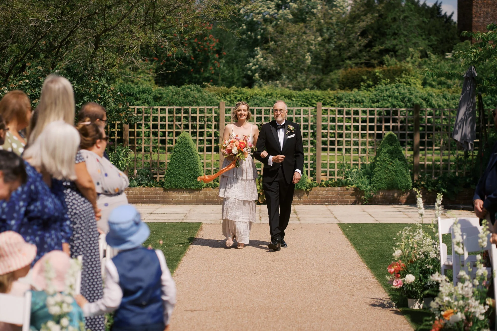 A bride walking down a garden aisle with her father between rows of guests at Hodsock Priory