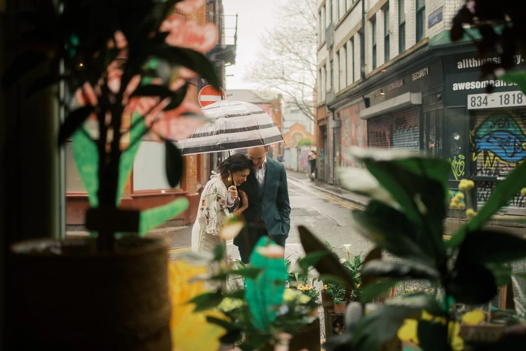 A couple laughing together under an umbrella on a rainy Manchester Northern Quarter street, seen through the plants in a shop window