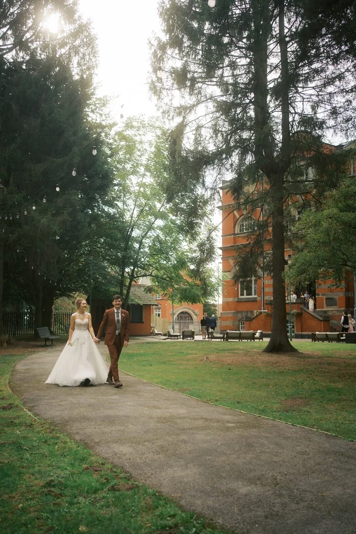 Bride and groom walking hand in hand along a curved path across the lawn at The Pumping House, with tall trees, strings of lights and the red brick building behind them.