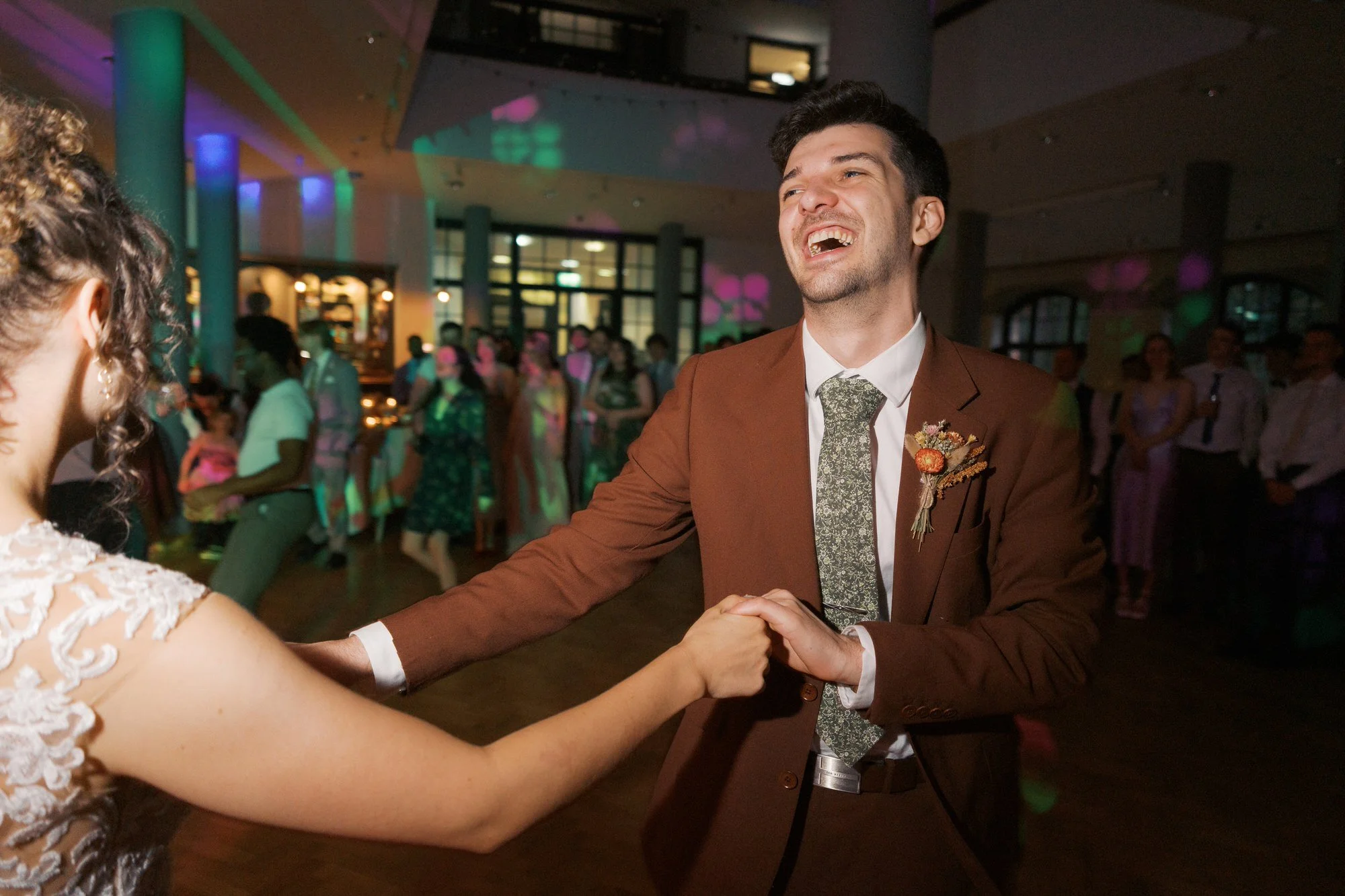 A groom laughing as he dances holding the bride's hands under coloured party lights, The Pumping House, Ollerton