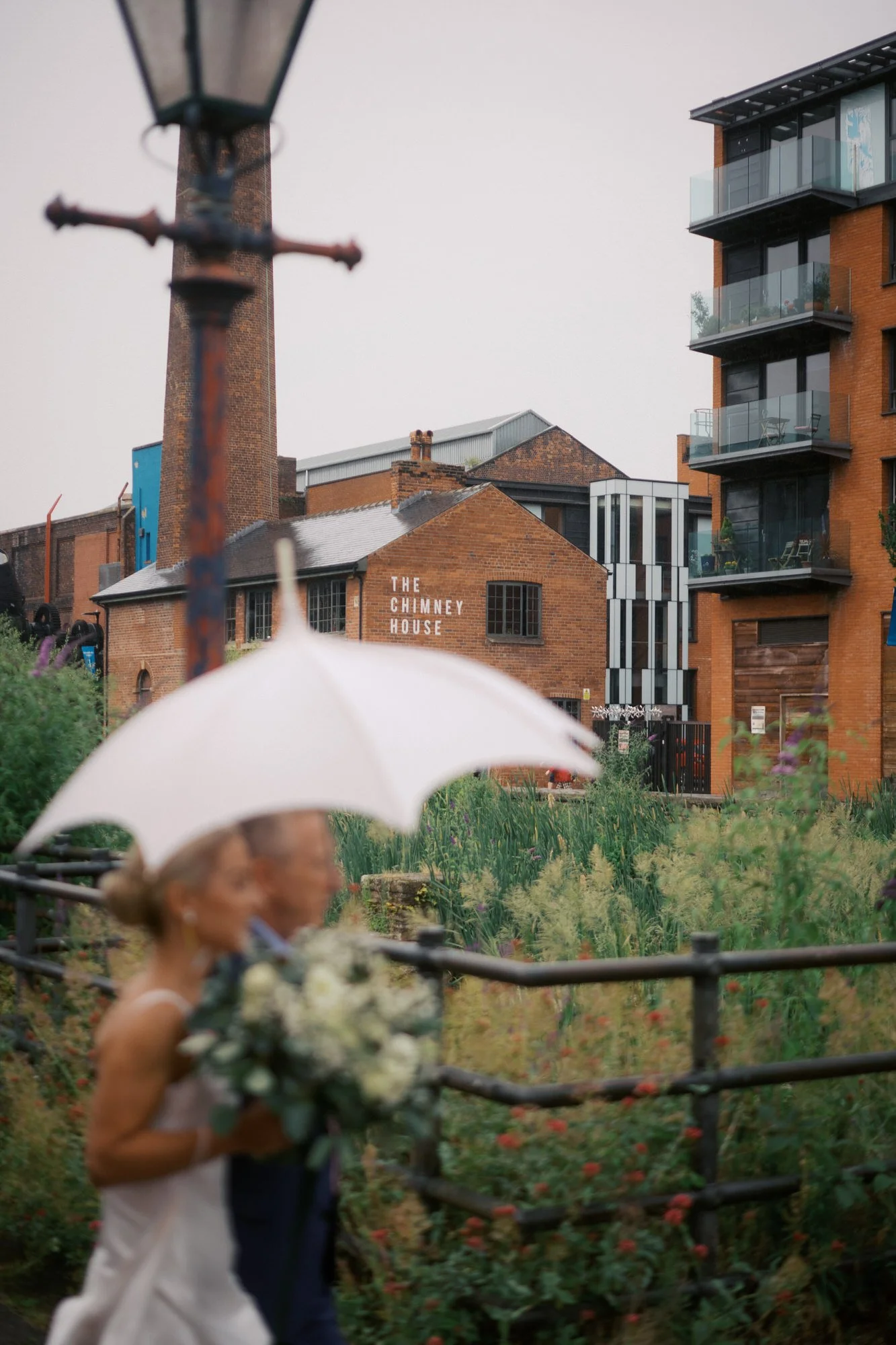 A bride with her bouquet walking under a white umbrella, with The Chimney House and its tall brick chimney behind her, Kelham Island, Sheffield