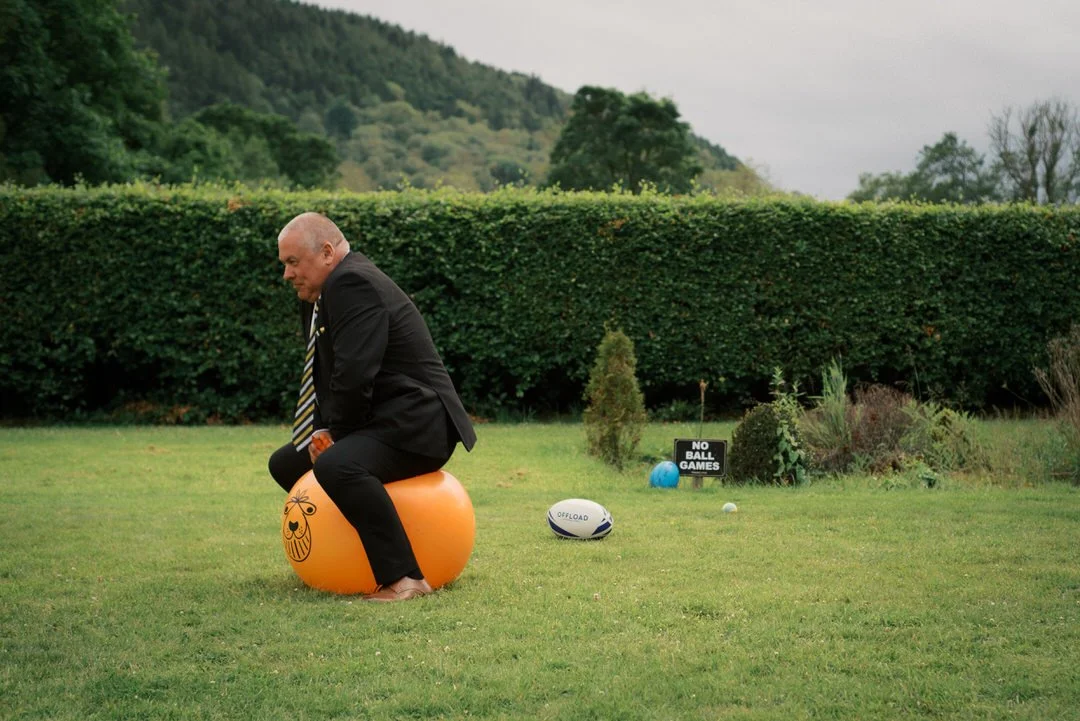 A man in a suit and striped tie sitting on an orange space hopper on a lawn at wedding at Hackness Grange, with a rugby ball on the grass and a "No ball games" sign behind him.