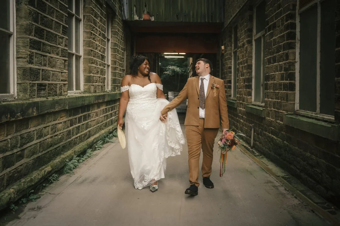 Bride and groom laughing and holding hands as they walk down a narrow lane between old stone buildings at The Venue in Halifax, the groom carrying her bouquet.