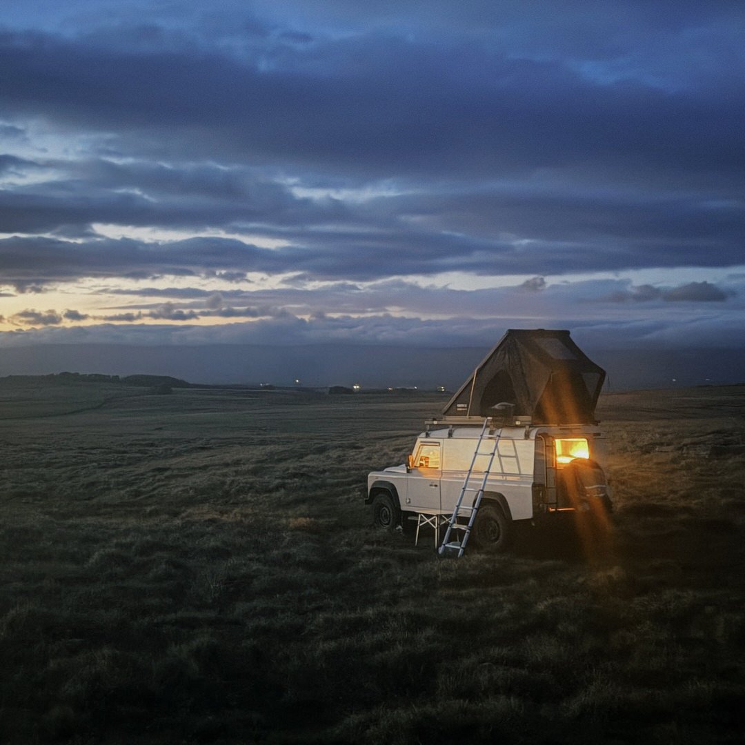 A white Land Rover in the Lake District with a tent open on its roof and a ladder leaning against its side, lit from inside, parked on open grassland at dusk under a cloudy sky.