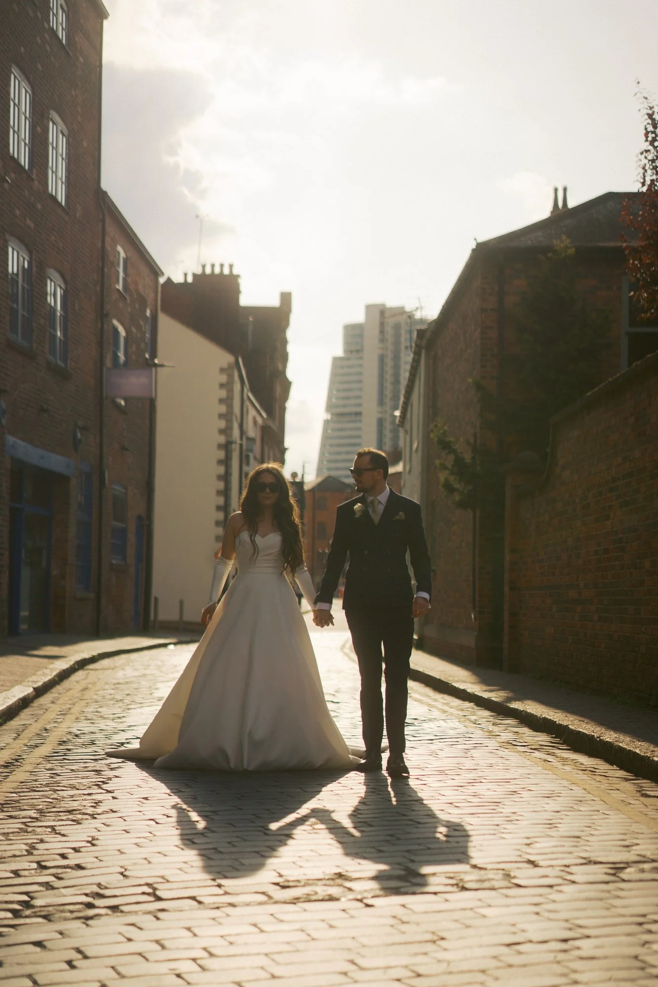 Bride and groom in sunglasses walking hand in hand down a sunlit cobbled street in Leeds, their shadows stretching towards the camera.