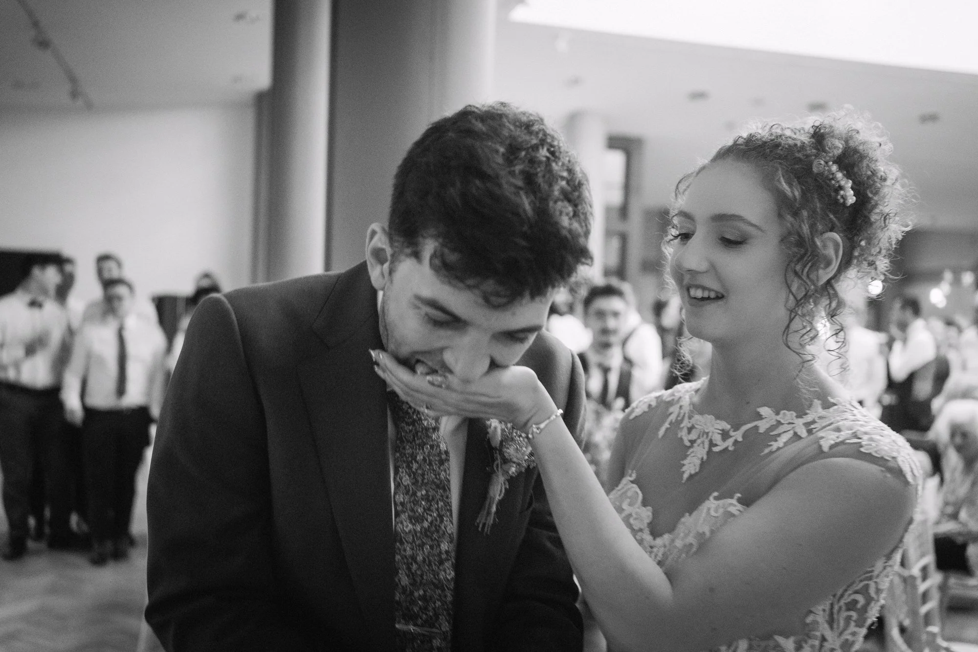 Black and white image of a bride holding her hand to her groom's mouth as guests look on, The Pumping House, Ollerton