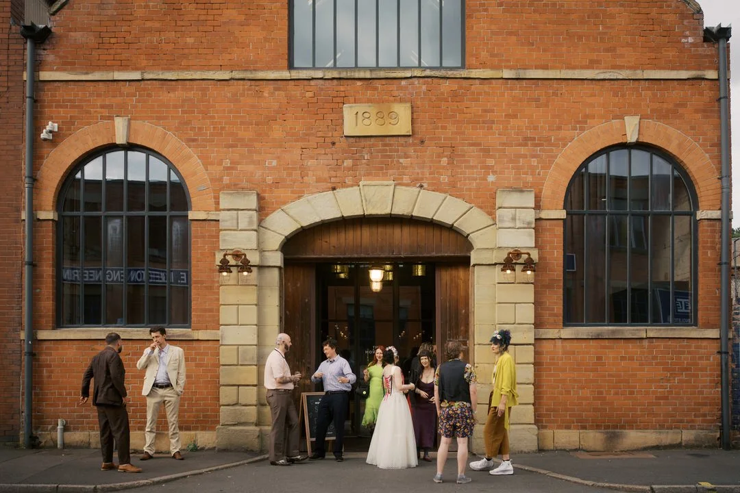 Bride in a flower headpiece and a dress laced with red ribbon chatting with guests outside the arched wooden doorway of The Mowbray, a red brick building with "1889" carved above the entrance.