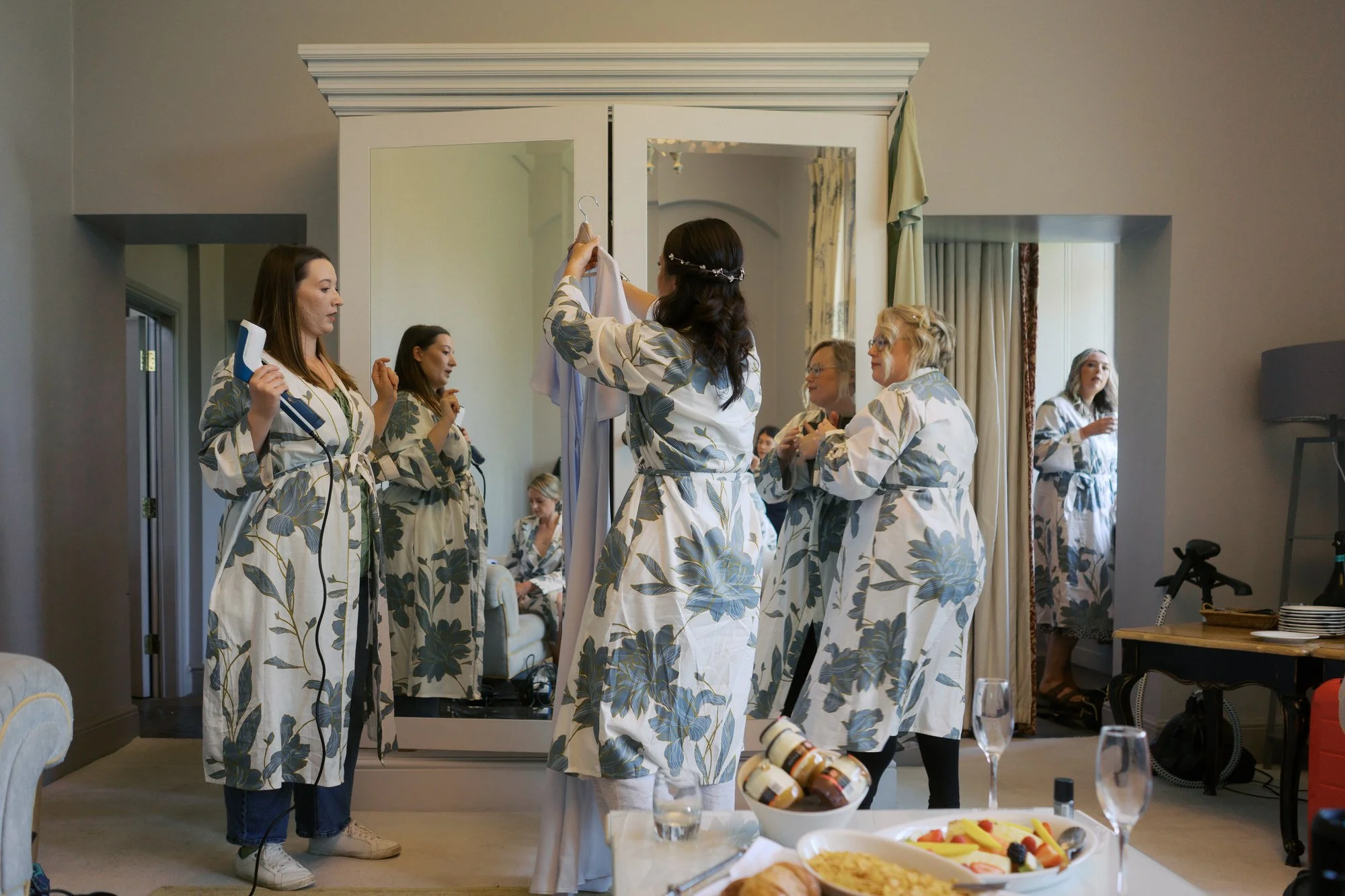 A bridal party in matching floral robes getting ready in front of a white mirrored wardrobe at Hodsock Priory