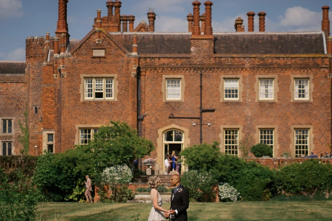 Bride and groom holding hands and facing each other on the lawn in front of the red brick house at Hodsock Priory, with guests gathered by the doorway behind them.
