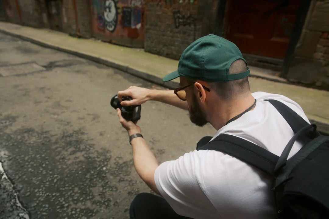 Wedding photographer in a green cap and backpack crouching in a narrow street in Manchester, holding a camera up to take a photo, seen from over his shoulder.