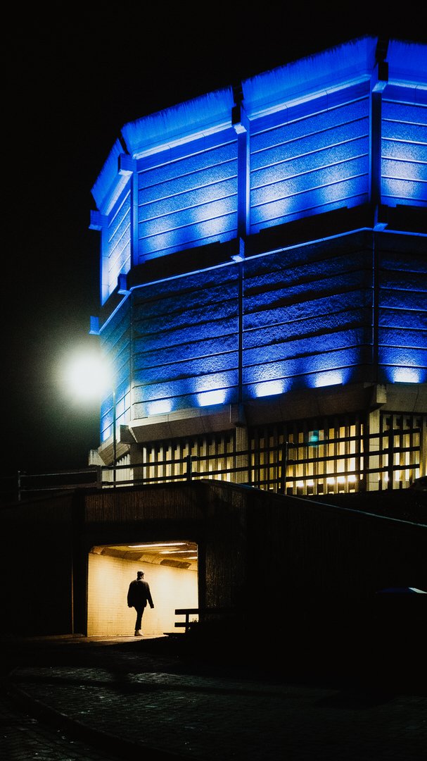 A street photograph of person walking alone into a lit underpass at night in Sheffield, beneath a large concrete building lit in blue.