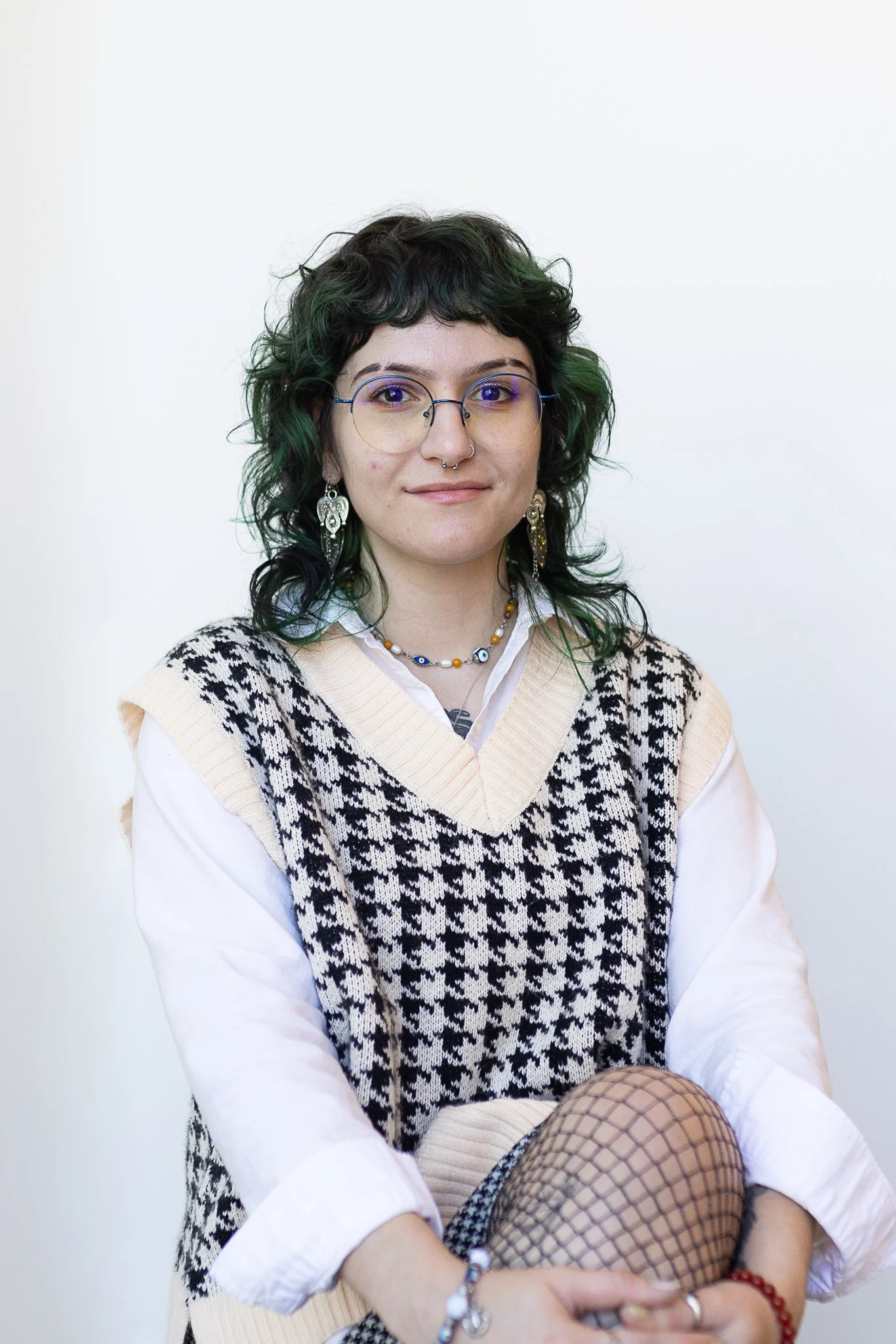 A stylist with shoulder-length curly green-tinted hair, wearing glasses and a houndstooth vest